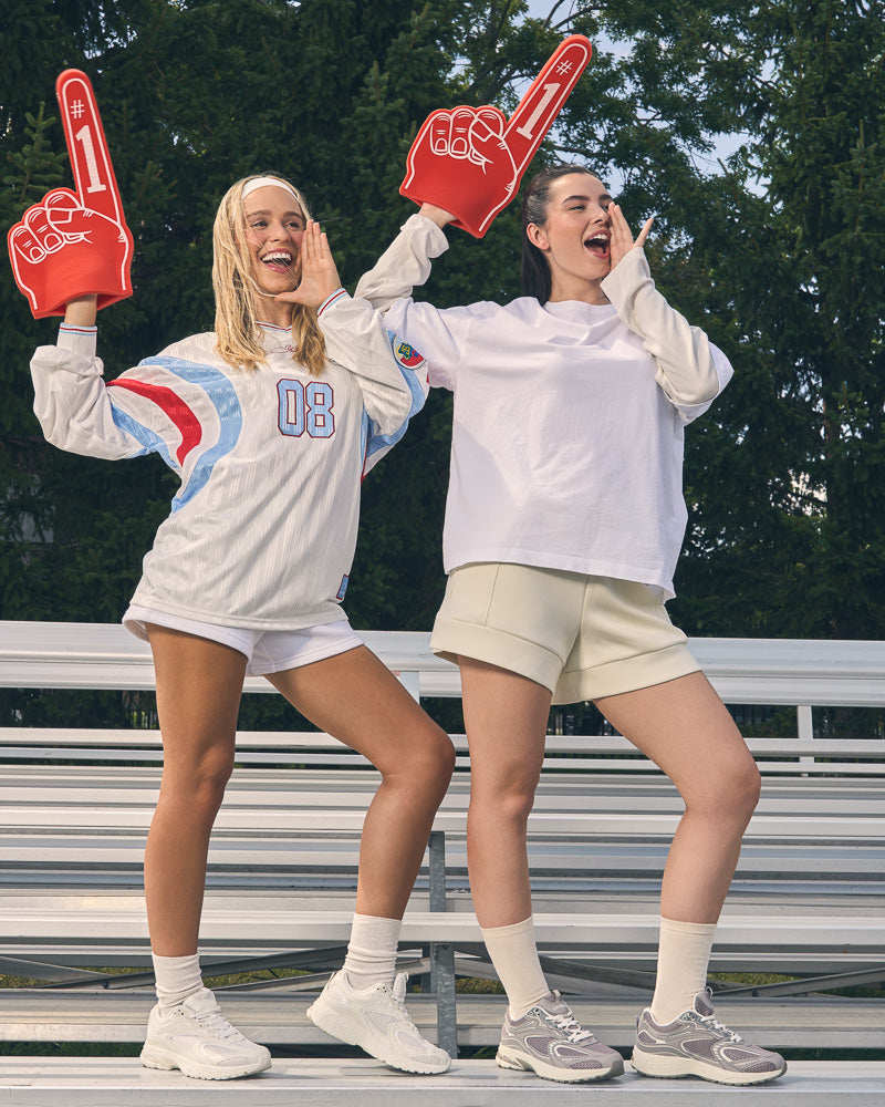 Yellow Shoes | Women's Athletic Shoes | CHARGE | Two women standing on outdoor bleachers wearing oversized white sweatshirts, shorts, and Yellow Shoes CHARGE sneakers, each holding a red foam finger. Trees and bleachers are visible in the background.