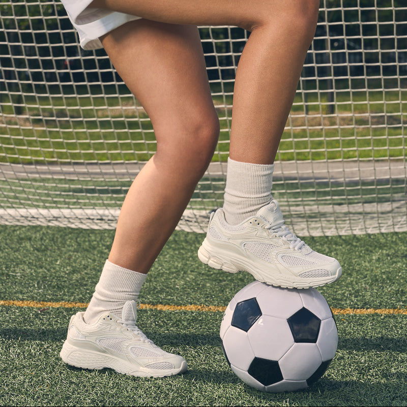 Yellow Shoes | Women's Athletic Shoes | CHARGE | A person in white athletic shorts and white sneakers kicks a black-and-white soccer ball on a green turf field in front of a white goal net.